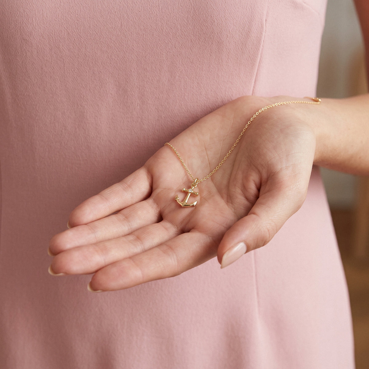 Small anchor pendant held in an open hand against a pink dress, showing its delicate scale