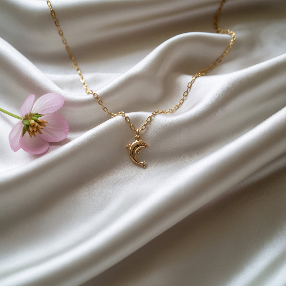 Dolphin pendant arranged on white silk with a pale pink cosmos flower beside the chain