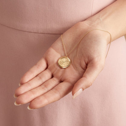 Gold watch face pendant cupped in a palm, showing the fluted edge and engraved dial