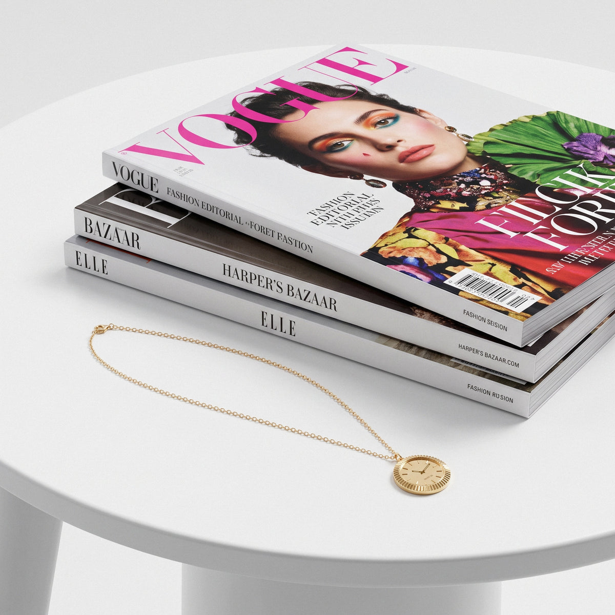 Watch face necklace coiled on a white side table beside a stack of fashion titles