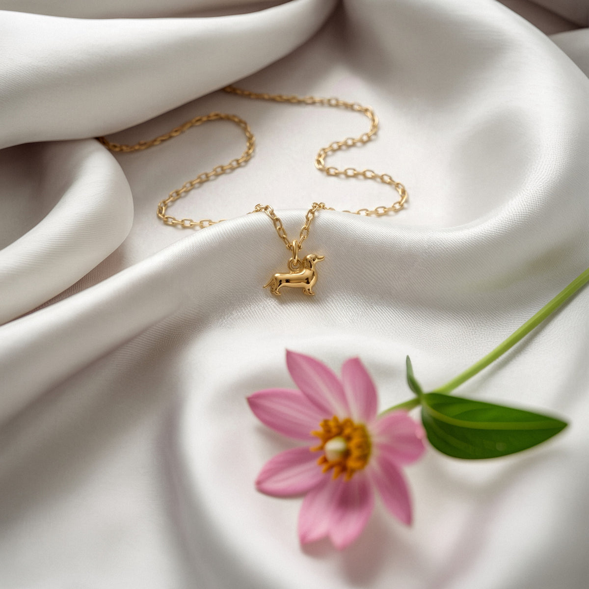Long dog pendant and chain arranged over white satin beside a pink cosmos flower