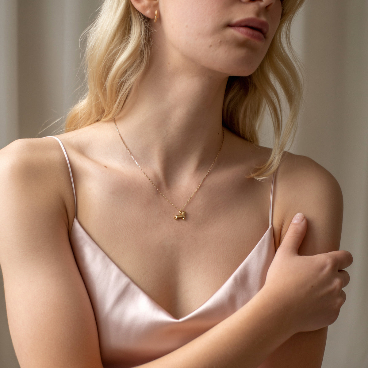 Tiny sausage dog necklace on a model in a satin camisole with one arm folded across her chest