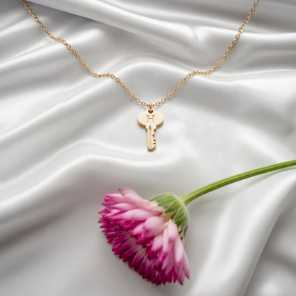Key initial necklace arranged on ivory silk beside a pink daisy, engraved letter facing up