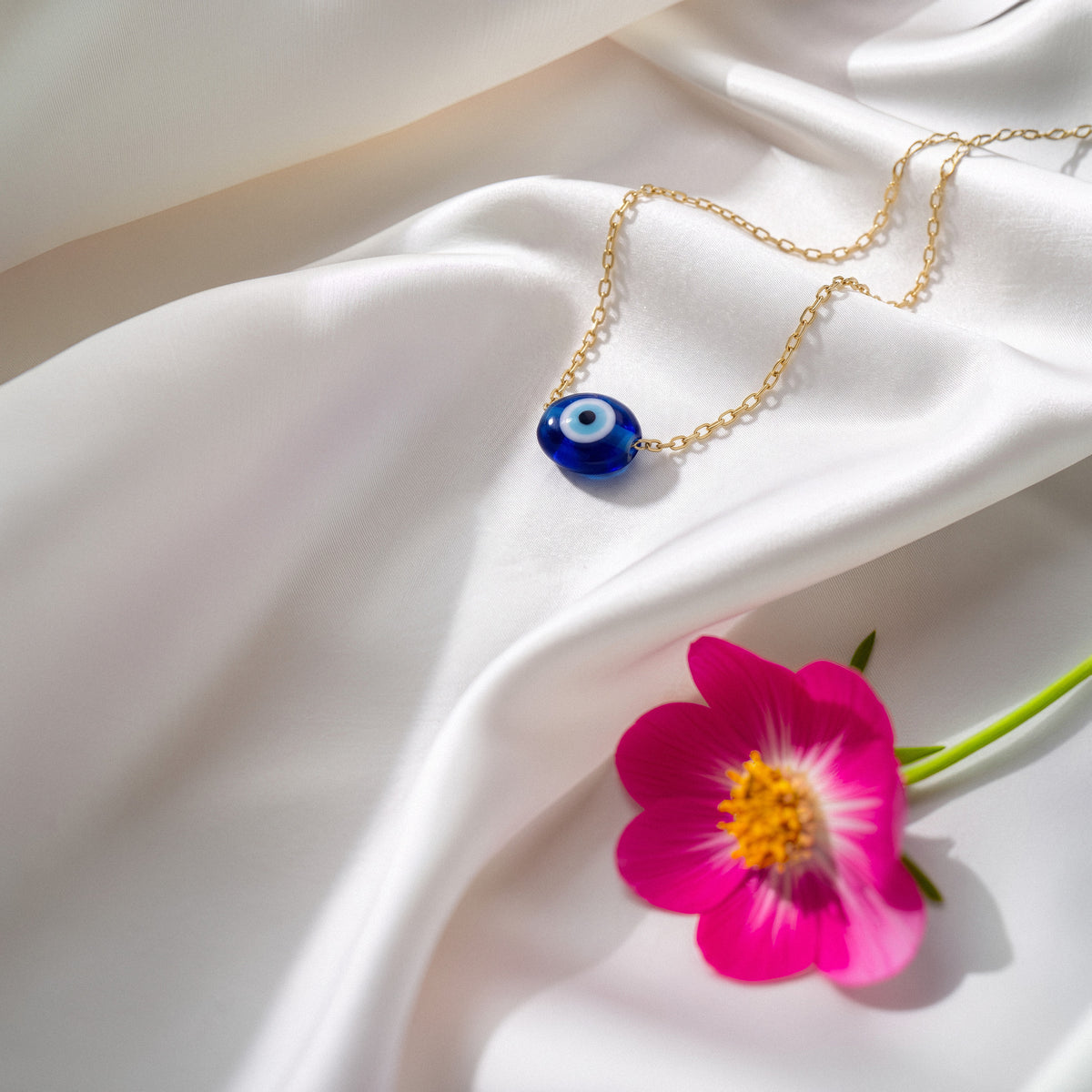 Cobalt evil eye necklace arranged on ivory silk beside a bright magenta flower