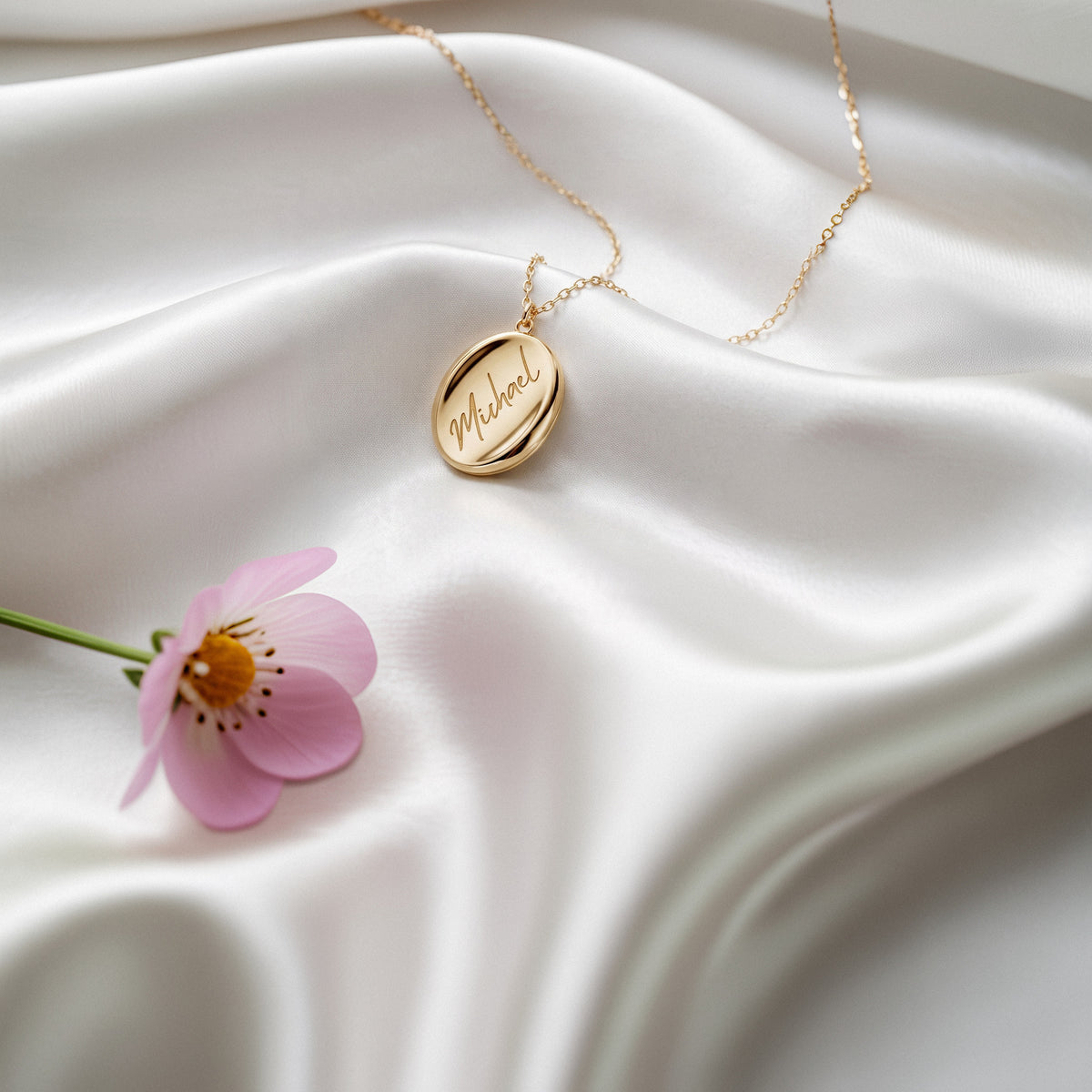 Oval name necklace on white silk beside a pink flower, script engraving visible