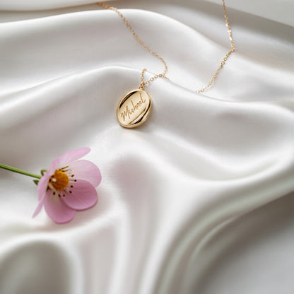 Oval name necklace on white silk beside a pink flower, script engraving visible
