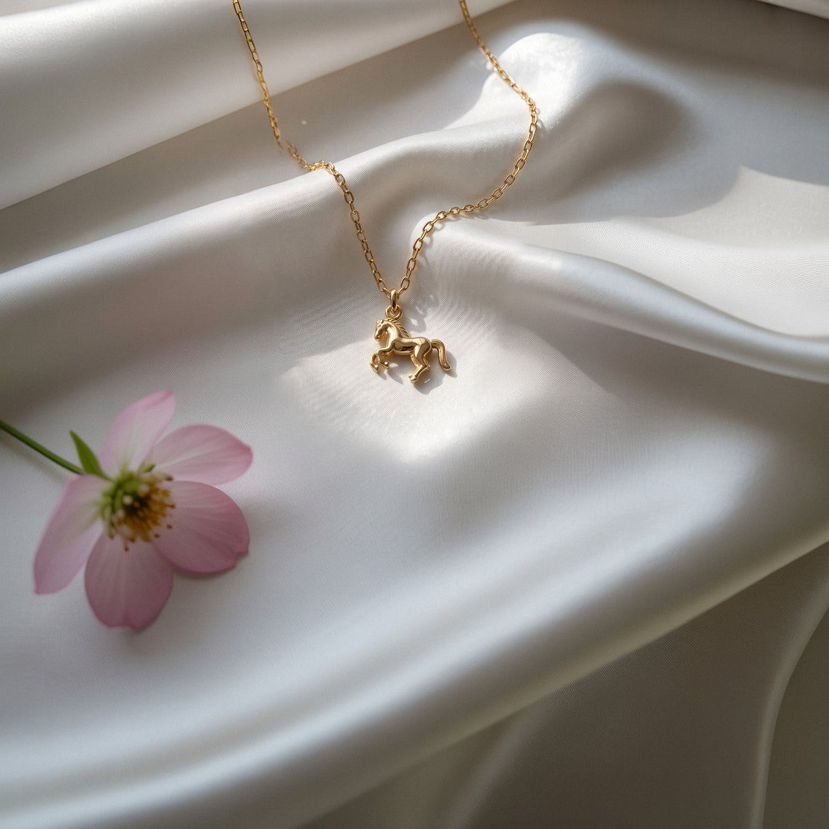 Rearing horse pendant catching light on folded ivory satin beside a pink cosmos