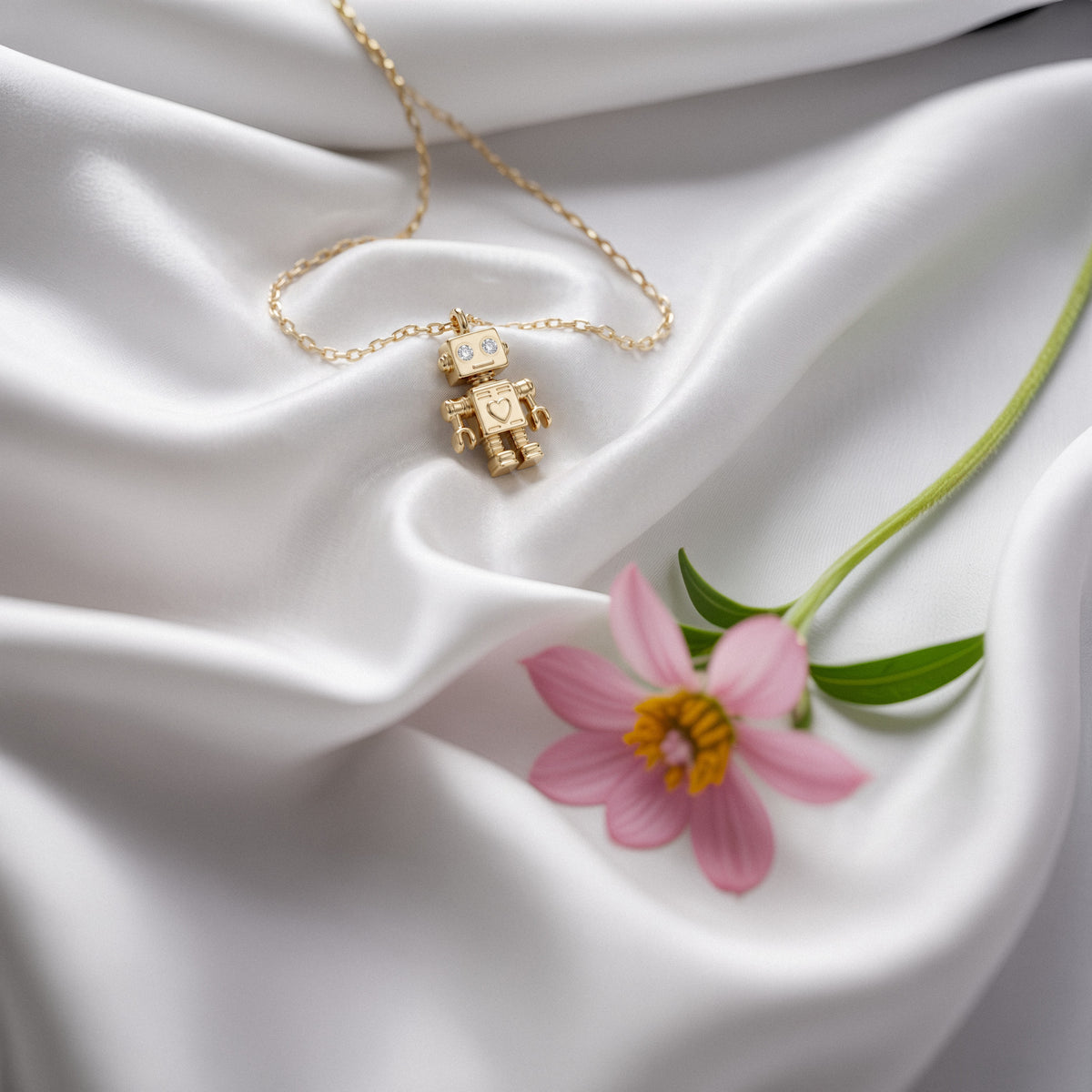 Robot necklace on white silk next to a pink cosmos flower, chain looped around the charm