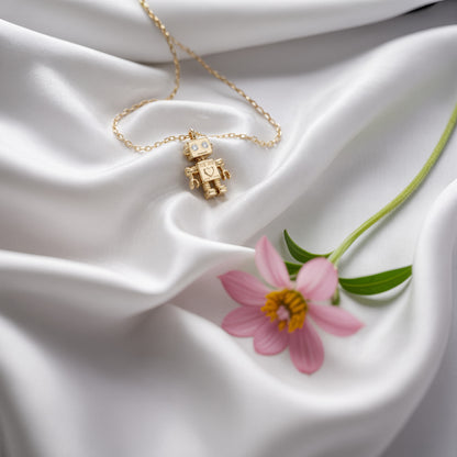 Robot necklace on white silk next to a pink cosmos flower, chain looped around the charm