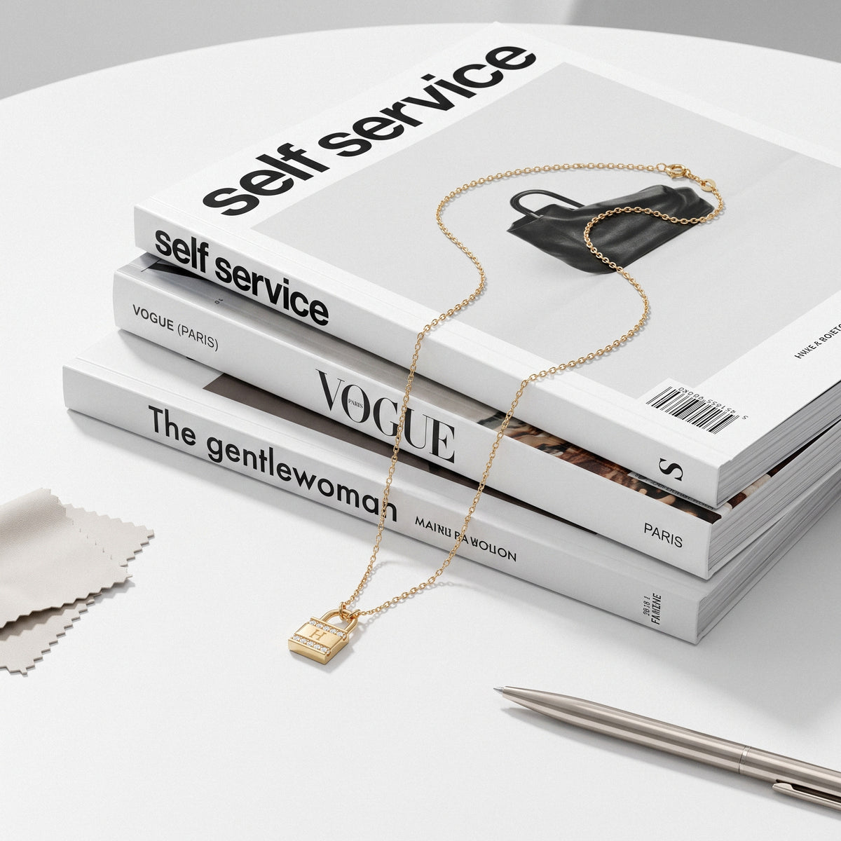Padlock initial necklace styled across Self Service and Vogue magazines on a white table