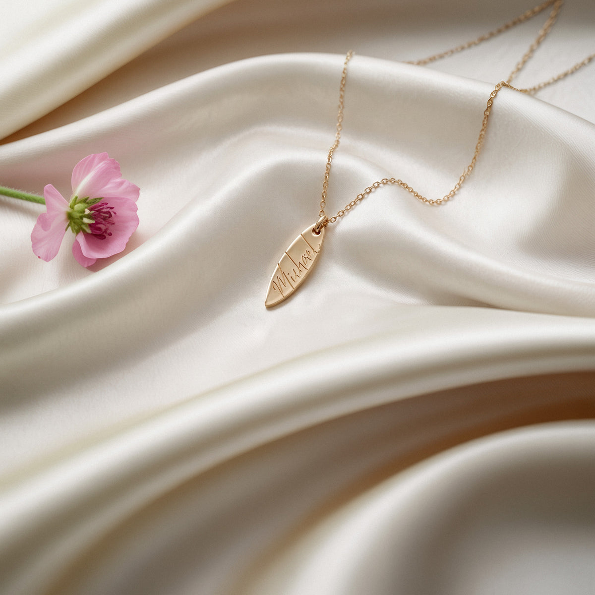 Personalised surfboard necklace on cream silk beside a pink poppy, engraving facing up