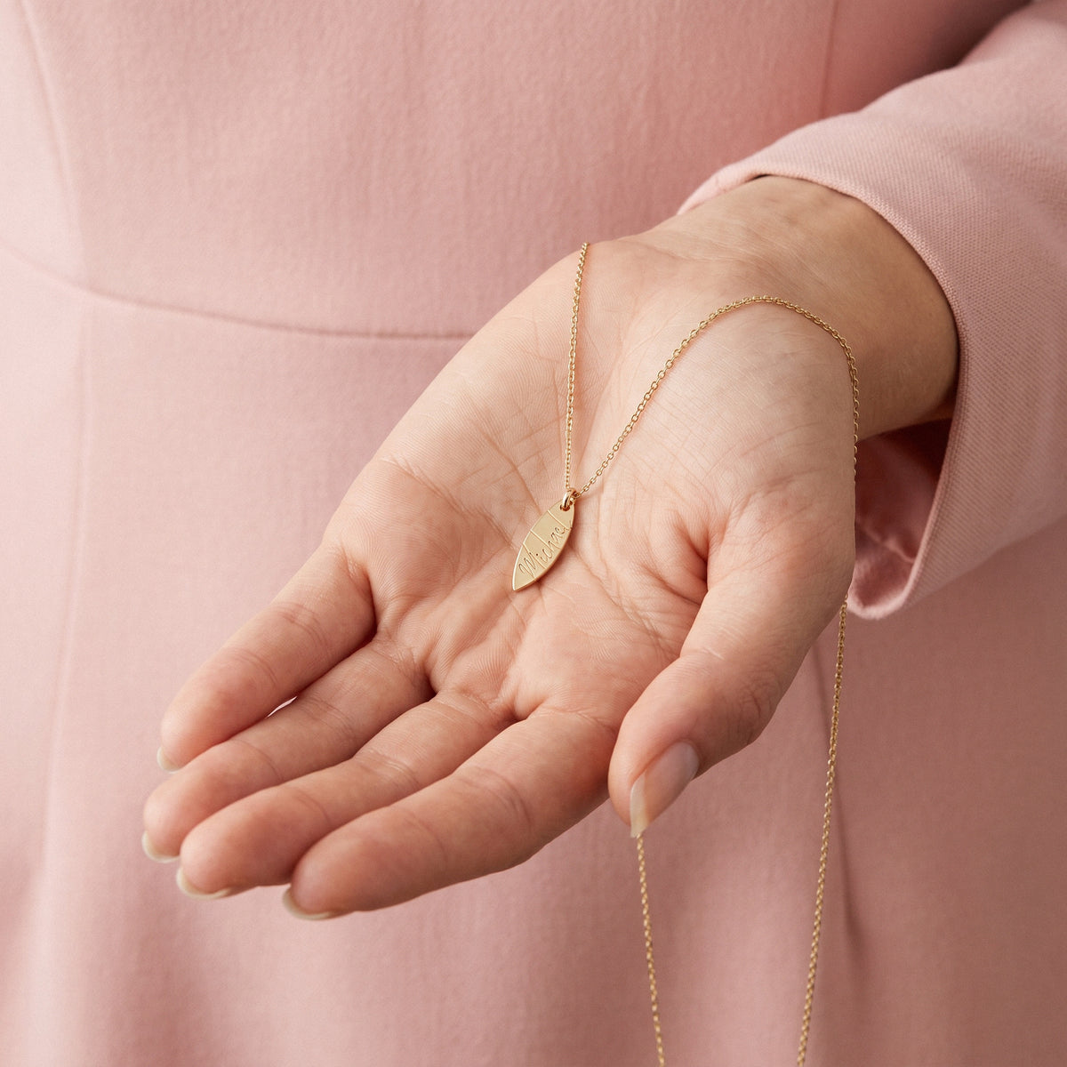 Name engraved surfboard pendant held in an open palm against a pink sleeve